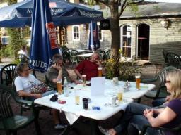 Fünf Personen sitzen an einem weißen Gartentisch mit Getränken im Freien unter blauen Sonnenschirmen vor einem hölzernen Gebäude