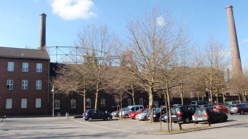 Parkplatz vor einem alten Fabrikgebäude mit zwei hohen Schornsteinen und kahlen Bäumen
