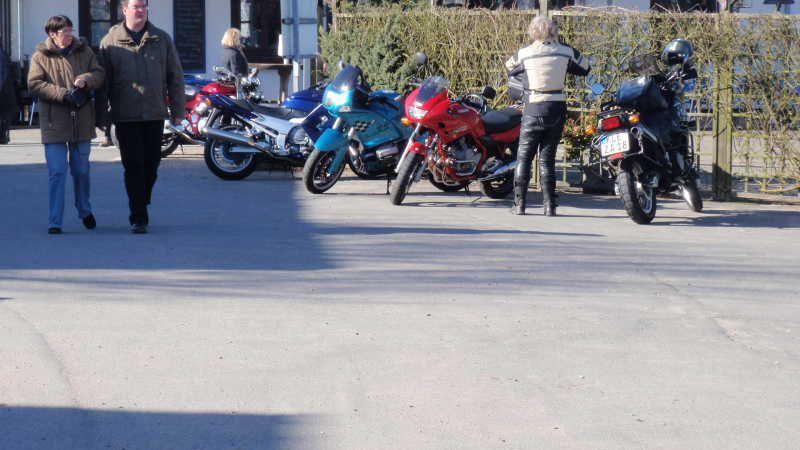 Mehrere Motorräder stehen geparkt auf einem Parkplatz, daneben spazieren zwei Personen.