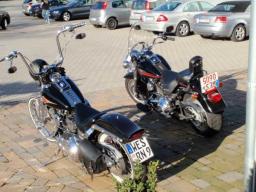 Zwei geparkte schwarze Motorräder auf gepflastertem Boden mit Pkw im Hintergrund