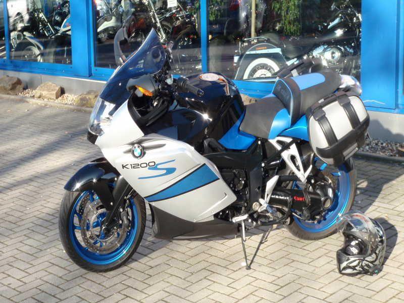 BMW K1200S Motorrad mit blauen und weißen Farben, geparkt vor einem blauen Gebäude mit Seitentaschen und Helm am Boden