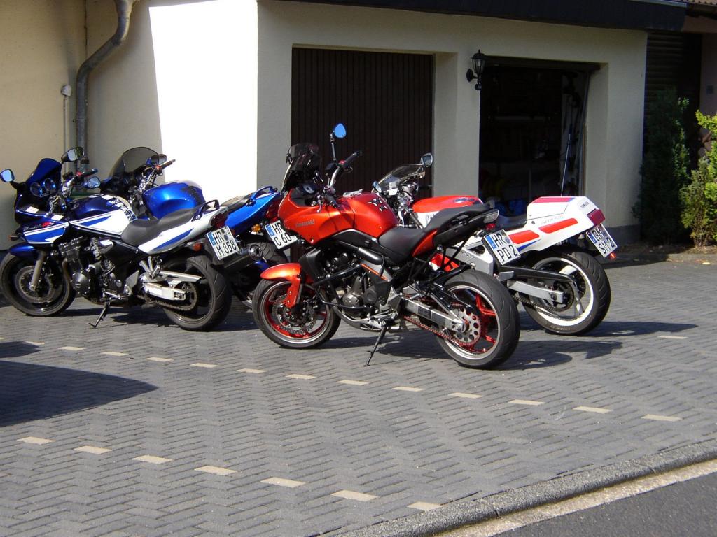 Vier Motorräder in verschiedenen Farben stehen geparkt vor einer Garage auf gepflastertem Untergrund.