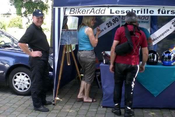 Drei Personen stehen vor einem Verkaufsstand mit dem Schild BikerAdd Lesebrille fürs Visier