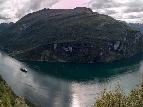 Schmaler Fjord mit bewaldeten Bergen und einem Schiff auf dem Wasser bei bewölktem Himmel