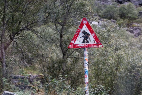 Verwittertes Verkehrszeichen mit einem Troll-Symbol in einer bewaldeten Landschaft