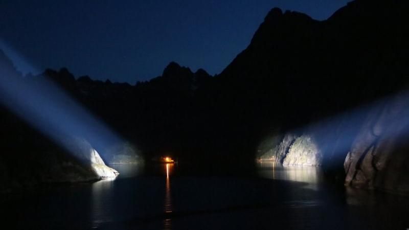 Landschaft mit See und Felsen bei Nacht, beleuchtet von starken Strahlern und einem kleinen Licht im Hintergrund