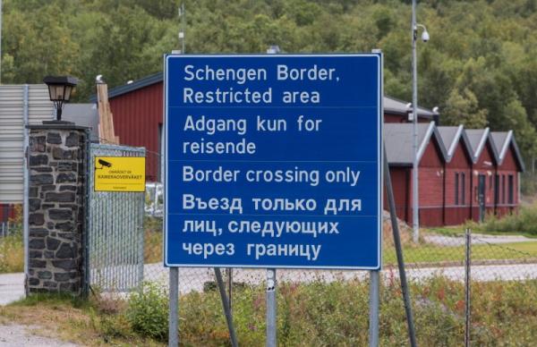 Blaues Grenzschild mit Warnungen in mehreren Sprachen vor einer geschlossenen Grenze im ländlichen Gebiet mit roten Gebäuden im Hintergrund