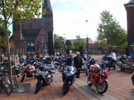Viele Motorräder und einige Menschen auf einem belebten Platz mit Gebäuden und Bäumen im Hintergrund