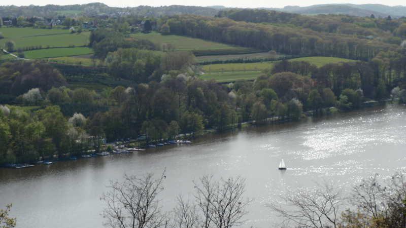 Fluss mit Segelboot vor bewaldeten Hügeln und grünen Feldern im Hintergrund