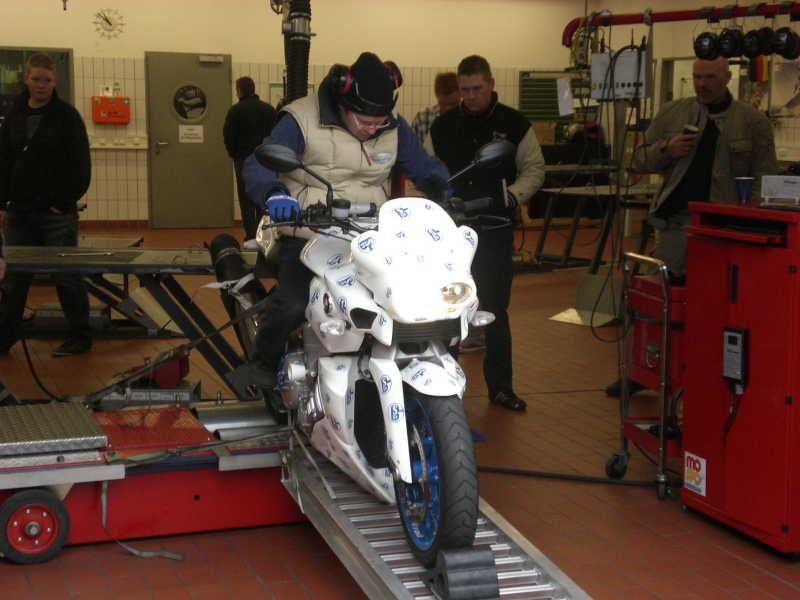 Person testet ein weißes Motorrad auf einem Prüfstand in einer Werkstatt mit mehreren Zuschauern