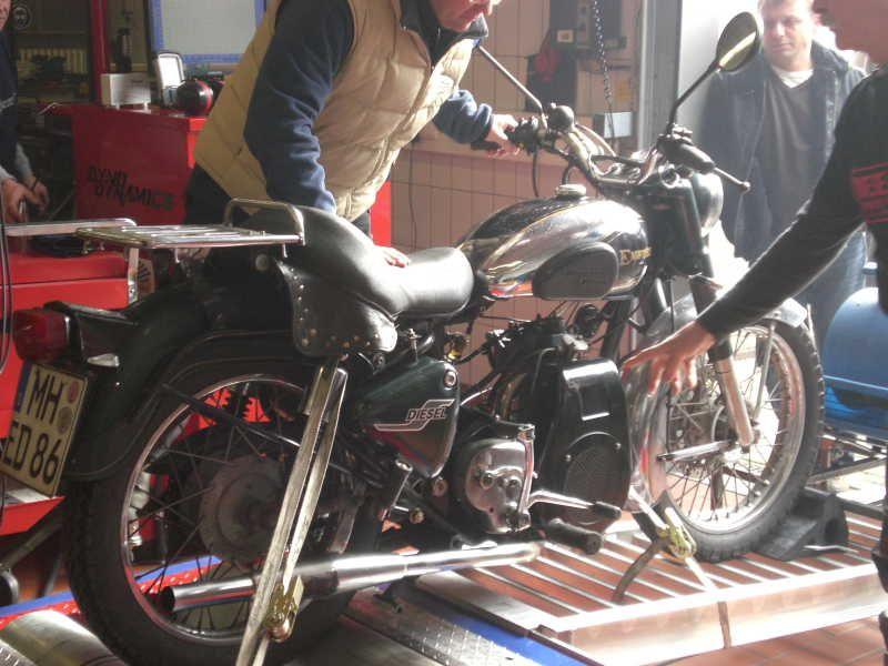 Personen begutachten ein auf einer Prüfrolle stehendes Motorrad in einer Werkstatt.
