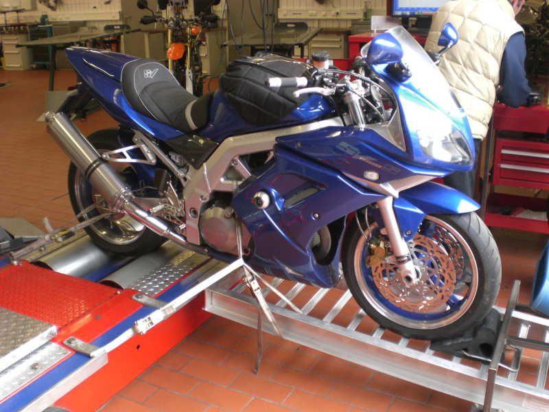 Blaues Motorrad auf Prüfstand in Werkstatt mit Person im Hintergrund