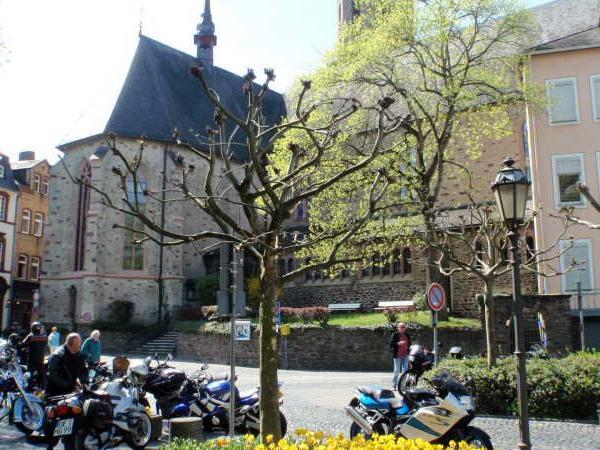 Blick auf einen Kirchplatz mit Motorrädern, Bäumen und historischen Gebäuden bei Sonnenschein