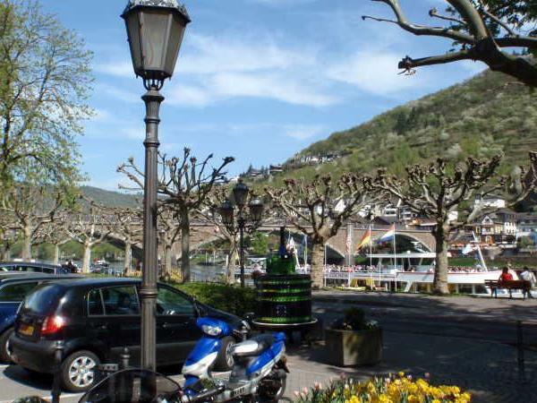 Parkplatz mit parkenden Autos und Motorroller, Baumreihe und hügelige Landschaft im Hintergrund