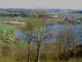 Landschaft mit einem See, hügeligen grünen Wiesen und einem kleinen Dorf am Ufer