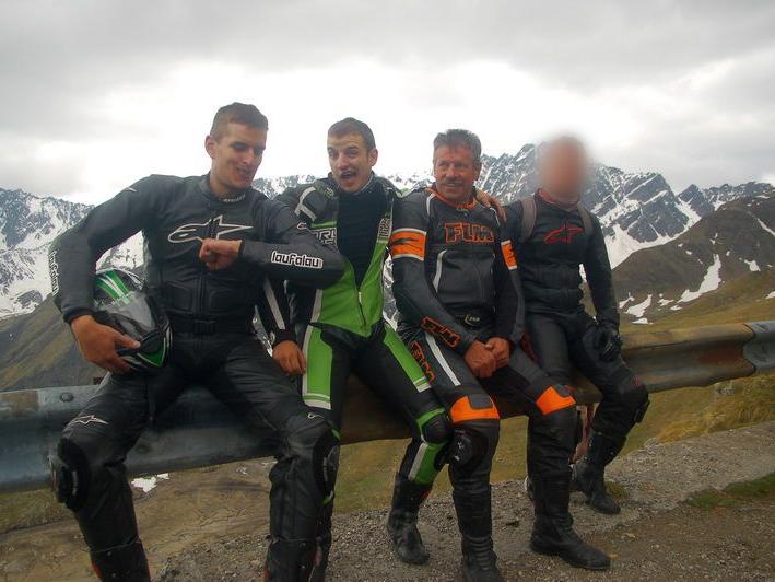 Vier Motorradfahrer in Schutzkleidung sitzen auf einer Leitplanke vor schneebedeckten Berggipfeln