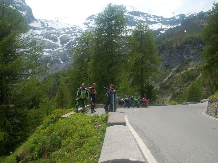 Drei Motorradfahrer stehen am Straßenrand vor einer Berglandschaft mit Schneegipfeln und grünen Bäumen