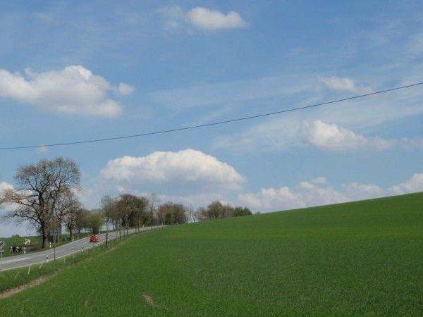 Landschaft mit grüner Wiese, Bäumen und einer Straße unter blauem Himmel mit Wolken