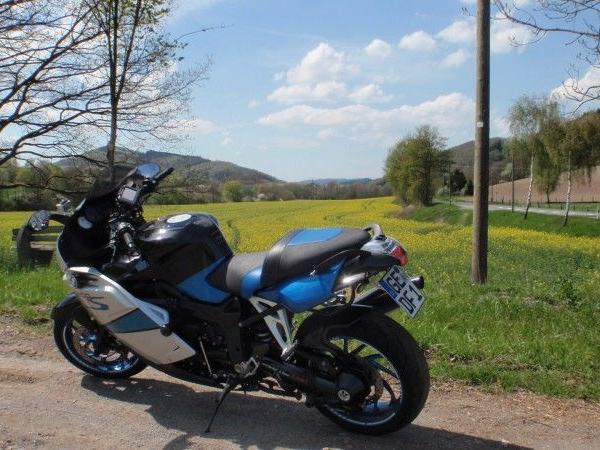 Schwarzes und blaues Motorrad steht auf einem Feldweg vor grüner Landschaft mit Bäumen und blühenden Wiesen