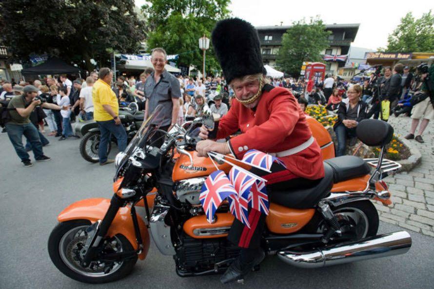 Mann in britischer Wachuniform auf oranger Motorrad mit Union-Jack-Fahnen bei einer Menschenmenge