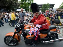 Mann in britischer Wachuniform auf oranger Motorrad mit Union-Jack-Fahnen bei einer Menschenmenge