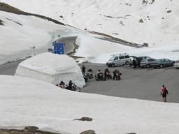 Parkplatz in einer verschneiten Berglandschaft mit Motorrädern, Autos und einer großen Schneehöhle