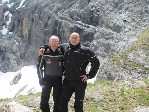 Zwei Männer in Motorradkleidung umarmen sich vor felsigem Bergpanorama mit Schneeflecken