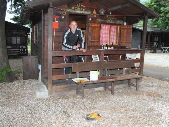 Mann lehnt an der Veranda eines kleinen Holzhauses mit rotem Vorhang und Bänken davor