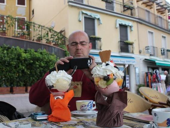 Mann fotografiert zwei bunte Eisschalen mit Früchten und Sahne in einem Straßencafé